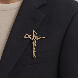 Puede incluir: Un broche de crucifijo dorado, que representa a Jesús en la cruz, prendido a una blazer azul marino oscuro. El diseño del broche es detallado y elegante. El color dorado contrasta con la tela oscura, lo que lo hace adecuado para ocasiones formales.