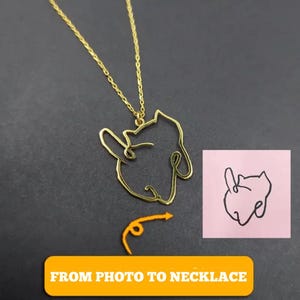 Op de afbeelding: Een goudkleurige ketting met een hanger in de vorm van een hondenkop, ontworpen als een doorlopende lijn. Een klein roze vierkant toont een zwarte lijntekening van de hondenkop. De tekst "FROM PHOTO TO NECKLACE" staat in een gele rechthoek.
