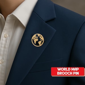 May include: A gold-colored world map brooch pin is pinned to a navy blue blazer. The pin is circular with a cut-out design of the continents. The text "WORLD MAP BROOCH PIN" is in a red rectangle.