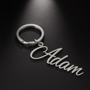 May include: Silver-colored keychain with the name "Adam" in a cursive font. The keychain has a circular ring attached to the name. The background is a dark gray color.