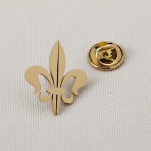 May include: A gold-colored fleur-de-lis pin and a matching pin back. The fleur-de-lis is a stylized lily, a symbol of French royalty. The pin is likely made of metal and has a smooth, polished finish. The pin back is round and has a clasp.