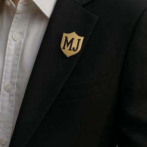 Op de afbeelding: Een goudkleurige, schildvormige reversspeld met de initialen "MJ" in een schreefletter is bevestigd aan een zwarte blazer. De blazer wordt over een wit overhemd gedragen. De speld is een decoratief accessoire.