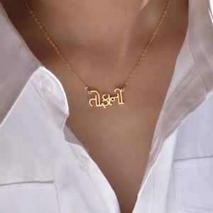 May include: A gold-colored necklace with a pendant featuring text in a script font. The necklace is worn against a white shirt. The chain is delicate and the pendant is centered.