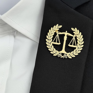 May include: A gold-colored lapel pin featuring the scales of justice, surrounded by a laurel wreath. The pin is affixed to a black blazer, with a white collared shirt visible in the background. The design is a symbol of law and justice.
