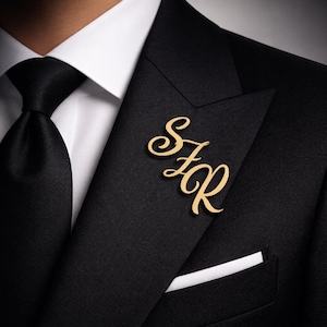 Op de afbeelding: Een goudkleurige monogramspeld met de initialen SFR, op een zwart colbert. De speld is in een sierlijk lettertype en is bij de revers geplaatst. Een zwarte stropdas en een wit pochet maken de formele outfit compleet.