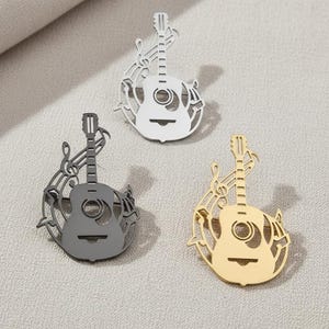 以下が含まれることがあります： 銀、ガンメタル、金の3つのギター型ブローチ。各ブローチは、音符が付いたギターのカットアウトデザインが特徴です。ブローチは、明るい灰色の表面に表示されています。