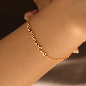 Puede incluir: Una pulsera dorada con los nombres "Nate", "Luna" y "Cael" en cursiva. La pulsera se lleva en la muñeca y la cadena es fina y delicada.