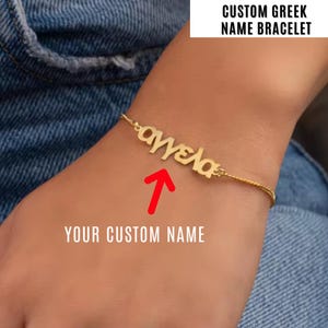 May include: A gold-colored custom Greek name bracelet. The bracelet features the Greek name "Αγγελα" in a stylized font. The text "CUSTOM GREEK NAME BRACELET" is displayed above the bracelet. The image also includes the text "YOUR CUSTOM NAME".