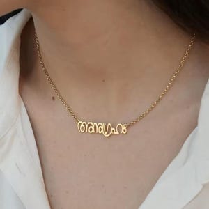May include: A gold necklace with a fine chain. The pendant displays a stylized inscription in a flowing script. The necklace is worn against a white shirt.
