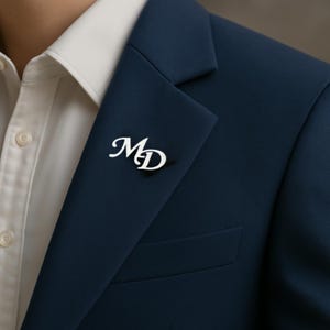 以下が含まれることがあります： ネイビーブルーのブレザーに、筆記体で「MD」のイニシャルが入ったシルバーカラーのラペルピンが付いています。ブレザーは白いシャツの上に着用されています。このピンは、フォーマルな装いにエレガントさを添えています。