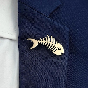 Peut inclure: Une épingle de revers dorée en forme d'arête de poisson. L'épingle est fixée à un blazer bleu marine. Le design de l'arête de poisson est détaillé et stylisé, avec une queue incurvée et un petit œil.