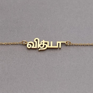 Peut inclure: Un collier doré avec le nom "வித்யா" écrit dans une police stylisée. Le nom est au centre, avec une chaîne délicate de chaque côté. Le fond est gris neutre.