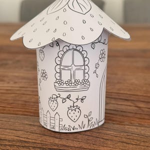 Puede incluir: Una casa de papel blanca con tem&aacute;tica de fresas. La casa presenta un techo festoneado, una ventana con jardineras, una puerta e ilustraciones de fresas. El dise&ntilde;o est&aacute; delineado en negro, listo para colorear.