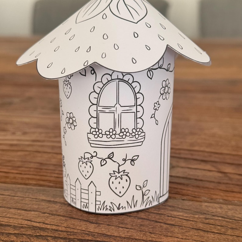 Puede incluir: Una casa de papel blanca con tem&aacute;tica de fresas. La casa presenta un techo festoneado, una ventana con jardineras, una puerta e ilustraciones de fresas. El dise&ntilde;o est&aacute; delineado en negro, listo para colorear.