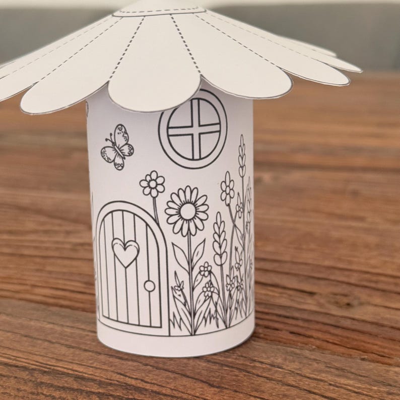 Puede incluir: Una casa de hadas de papel blanco con un techo en forma de margarita. La casa presenta una puerta con un coraz&oacute;n, una ventana redonda e ilustraciones de flores y una mariposa, todo delineado en negro.