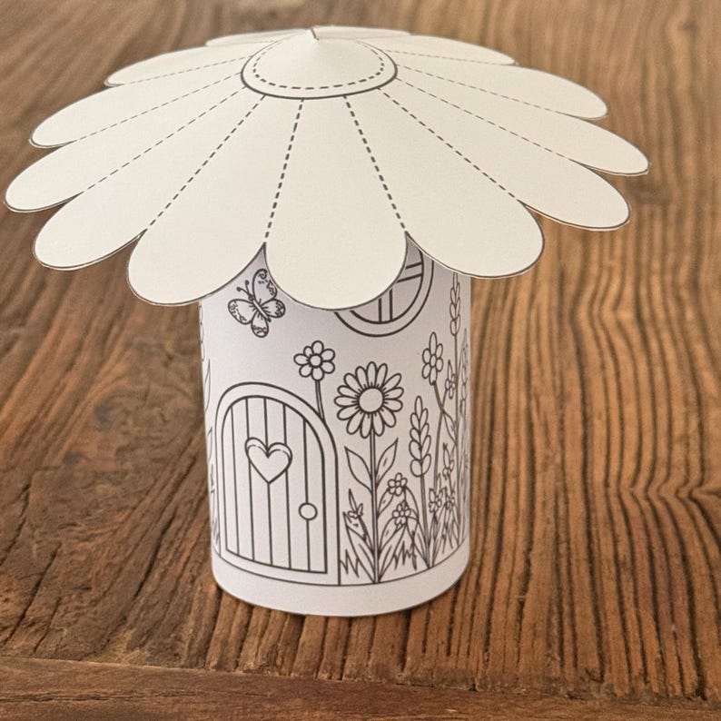 Puede incluir: Una manualidad de papel blanco de una casa de hadas con un techo en forma de margarita. El cuerpo cil&iacute;ndrico presenta una puerta con forma de coraz&oacute;n, una ventana e ilustraciones florales y de mariposas. El techo est&aacute; unido con una l&iacute;nea punteada.