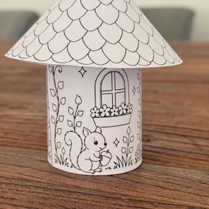 Puede incluir: Una manualidad en forma de hongo de papel blanco, con un techo festoneado y una base cil&iacute;ndrica. La base presenta una escena dibujada de una ardilla sosteniendo una bellota, una ventana con flores y enredaderas. Dise&ntilde;ado para colorear.