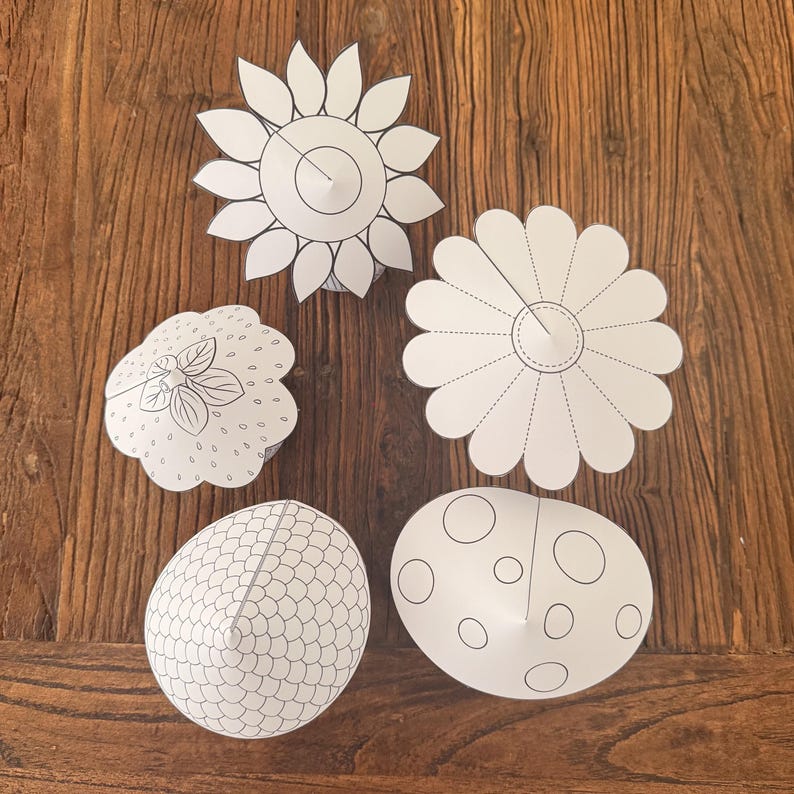 Puede incluir: Cinco recortes de papel blanco de varias formas, incluyendo un girasol, una margarita, una fresa y dos dise&ntilde;os en forma de huevo. Los recortes est&aacute;n sobre una superficie de madera oscura. Cada recorte tiene un contorno negro y detalles interiores.