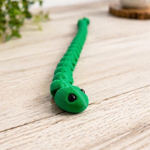May include: A vibrant green articulated snake toy with black eyes is positioned on a light wooden surface. The toy is composed of linked sections, enabling it to flex and move. The snake's head is rounded, and the body narrows to a point.