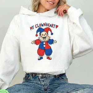 May include: A white hooded sweatshirt featuring a graphic of a clown wearing a red and blue outfit with a ruffled collar. Above the clown, the text reads MY CLOWNHEART in red and blue letters, accompanied by a small red heart symbol on the right side.