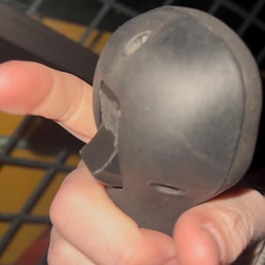 May include: A close-up of a black, rounded object with a rectangular protrusion and small holes. The object is held in a hand, with the fingers visible. The object appears to be made of a hard, possibly plastic material.