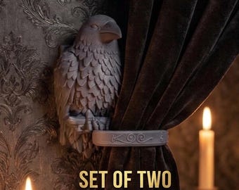 May include: A set of two gray curtain tiebacks featuring a sculpted raven figure with detailed plumage. The tieback band includes a decorative embossed design with the text KOC. The raven is shown mounted on a wall, holding a dark brown curtain.