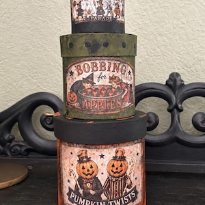 May include: A stack of three cylindrical boxes with Halloween-themed graphics. The top box is black with a Trick or Treat Parade label. The middle box is green with a Bobbing for Apples label. The bottom box is orange with a Pumpkin Twists label.