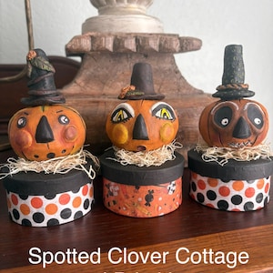 May include: Three hand-painted pumpkin figurines with painted faces and hats, each resting on a round box base. The bases feature orange, black, and white polka dot or candy patterns. The text Spotted Clover Cottage and Primitives is visible.