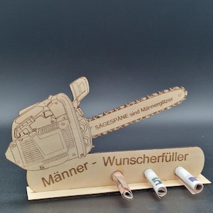 May include: A wooden money holder shaped like a chainsaw, featuring the text "SÄGESPÄNE sind Männerglitzer" and "Männer - Wunscherfüller". It has three holes holding rolled banknotes. The design is laser-cut with a natural wood finish.