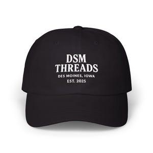 May include: Black baseball cap with the white text "DSM THREADS DES MOINES, IOWA EST. 2025" across the front. The cap has a curved brim and a fabric strap closure on the back.