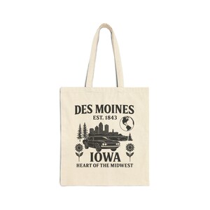 May include: Cream-colored tote bag featuring a black graphic design. The design includes the text "DES MOINES EST. 1843 IOWA HEART OF THE MIDWEST" along with a car, cityscape, trees, and sunflowers. The bag has two handles.