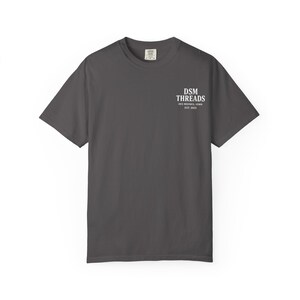 May include: A dark gray t-shirt with short sleeves. The shirt features the text "DSM THREADS DES MOINES, IOWA EST. 2023" printed in white on the upper right chest area. The shirt is made of a soft material.