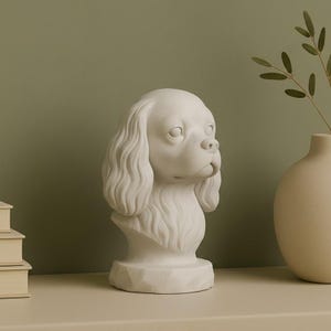 May include: A white dog bust sculpture with detailed fur and facial features. The sculpture is on a pedestal base. A vase with greenery is to the right, set against a neutral background.