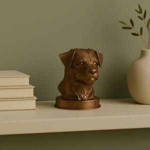 Op de afbeelding: Een bronzen hondenbuste met gedetailleerde vachttextuur. De hondenkop is gemonteerd op een rechthoekige basis. De sculptuur staat op een witte plank met een stapel boeken en een vaas.