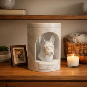 May include: A marble-patterned pet urn with a white French Bulldog bust inside an arched niche. The urn has a rectangular nameplate with the name "WALAGE" and the years "2013-2024". A framed photo of a French Bulldog and a lit candle are nearby.