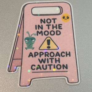 May include: A pink, glittery sticker in the shape of a caution sign. The text reads "NOT IN THE MOOD" above a yellow warning sign and "APPROACH WITH CAUTION" below. Includes a green alien and a yellow, frowning face.
