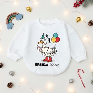 May include: White baby onesie with a cartoon goose wearing a party hat and red boots, holding a bottle and balloons. The text on the onesie reads "GRUMPY BIRTHDAY GOOSE". Festive holiday decorations surround the onesie.
