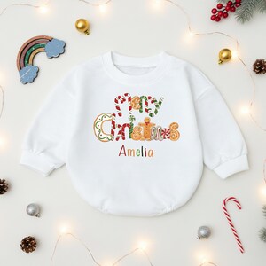 May include: White baby bodysuit with long sleeves. The words "Merry Christmas" are written in a gingerbread cookie design, with the name "Amelia" below. The bodysuit is decorated with a rainbow, ornaments, and Christmas lights.