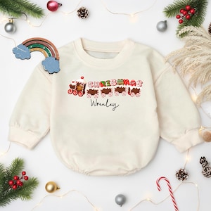 May include: Cream-colored baby romper with a Christmas train design. The train features gingerbread-style cars with the word "CHRISTMAS" and the name "Wrenley" printed in black. Festive holiday decor surrounds the romper.