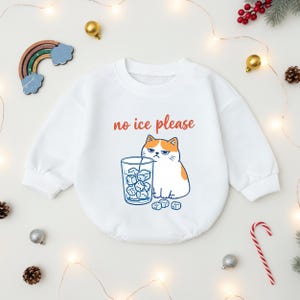 May include: White baby bodysuit with the phrase "no ice please" in red script. The graphic features a grumpy cartoon cat with orange and white fur, looking at a glass of ice cubes. The bodysuit is surrounded by holiday decorations.