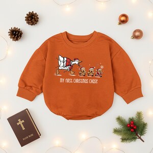 May include: An orange baby bodysuit with long sleeves. The front features a cartoon goose in a Santa suit chasing gingerbread men. The text "MY FIRST CHRISTMAS CHASE" is below the graphic. The bodysuit is surrounded by Christmas decorations.