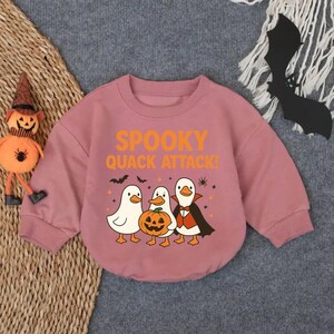 May include: Dusty rose-colored baby bodysuit with the text "Spooky Quack Attack!" and a graphic of three ducks dressed for Halloween. One duck holds a carved pumpkin. Black bats and stars are also featured.