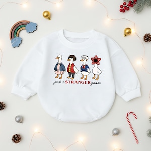 May include: White baby onesie with a graphic of four cartoon geese dressed as characters. The text "just a STRANGER goose" is printed below the geese in red and black. The onesie is surrounded by holiday decorations.