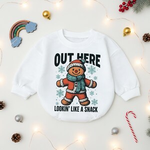 May include: White baby onesie with a gingerbread man graphic wearing a hat and scarf. The text "OUT HERE LOOKIN' LIKE A SNACK" is printed above and below the gingerbread man. The onesie is surrounded by Christmas decorations.