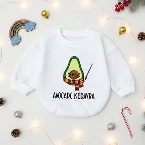 May include: White baby onesie with a cartoon avocado wearing glasses and a scarf, holding a wand. The text "AVOCADO KEDAVRA" is printed below the avocado illustration. Festive decorations surround the onesie.