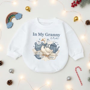May include: White baby onesie with the text "In My Granny era" and a graphic of a goose, blue teapot, and bowls. The onesie is surrounded by holiday decorations, including a rainbow, ornaments, and a candy cane.