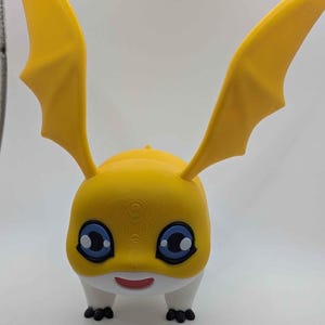 May include: A yellow figurine of a cartoon character with large, wing-like ears. The character has blue eyes, a white belly, and small black feet. The figurine is likely 3D-printed and stands on a white surface.