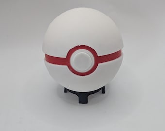 Puede incluir: Réplica de Poké Ball blanca y roja, un objeto esférico con una banda roja central y una base negra. La bola tiene un botón circular en el centro de la banda roja. El objeto es un artículo de colección o decorativo.
