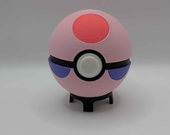 Puede incluir: Un juguete esférico con una mitad superior de color rosa claro con un círculo rojo en el centro, una banda horizontal negra y un botón circular blanco. La mitad inferior es rosa claro con dos acentos morados sobre un soporte negro.