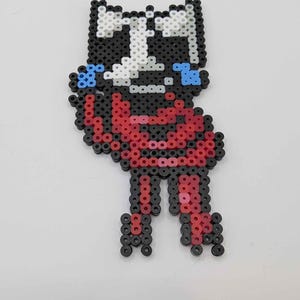 May include: A pixel art figure made from colorful plastic beads. The figure has a black and white head, red body, and black legs. Blue accents are on the figure's face. The figure is standing on a white surface.